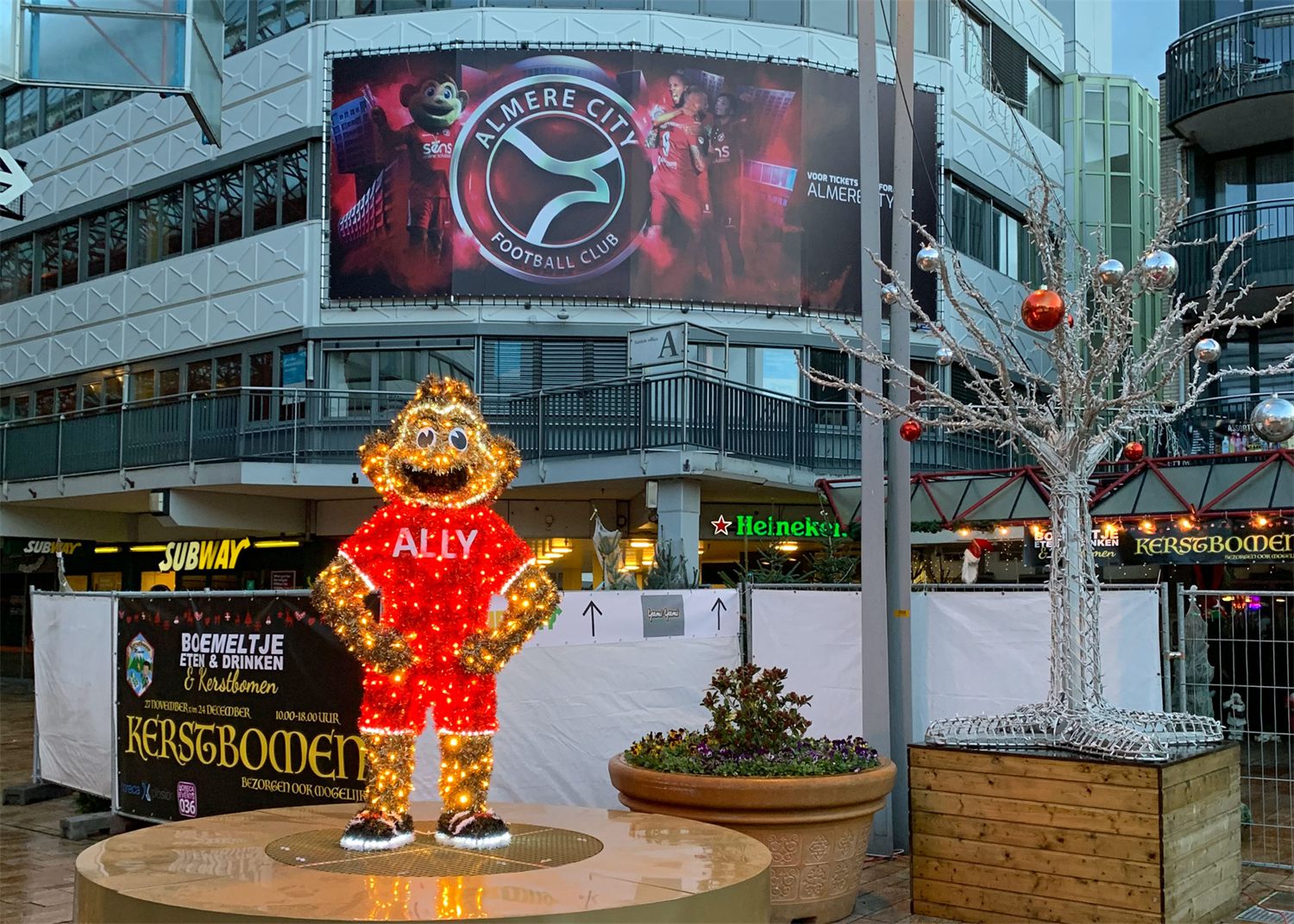 Verlichte mascotte in een rood shirt op een plein, naast een zilveren lichtboom