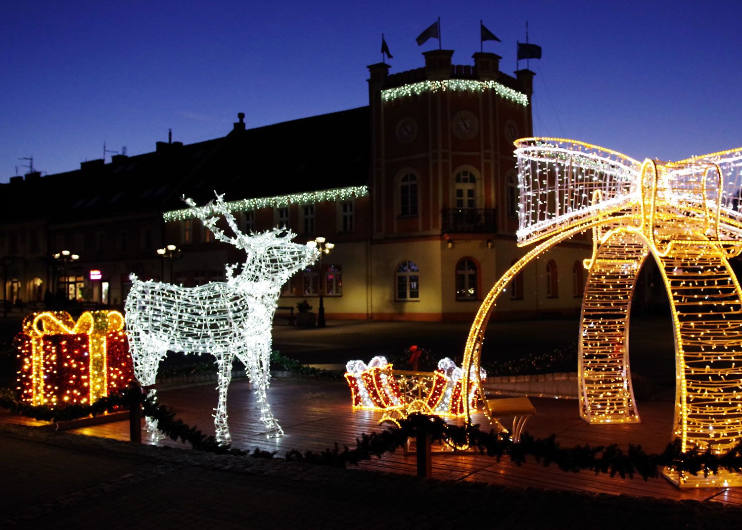 Verlicht rendier, cadeaus en lichtbogen op een plein bij avond