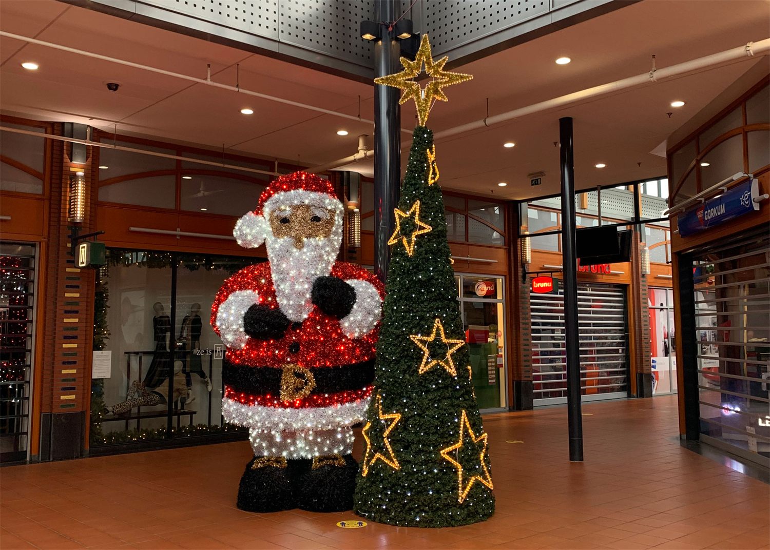 Verlichte kerstman naast een kegelvormige kerstboom met gouden sterren in een winkelcentrum