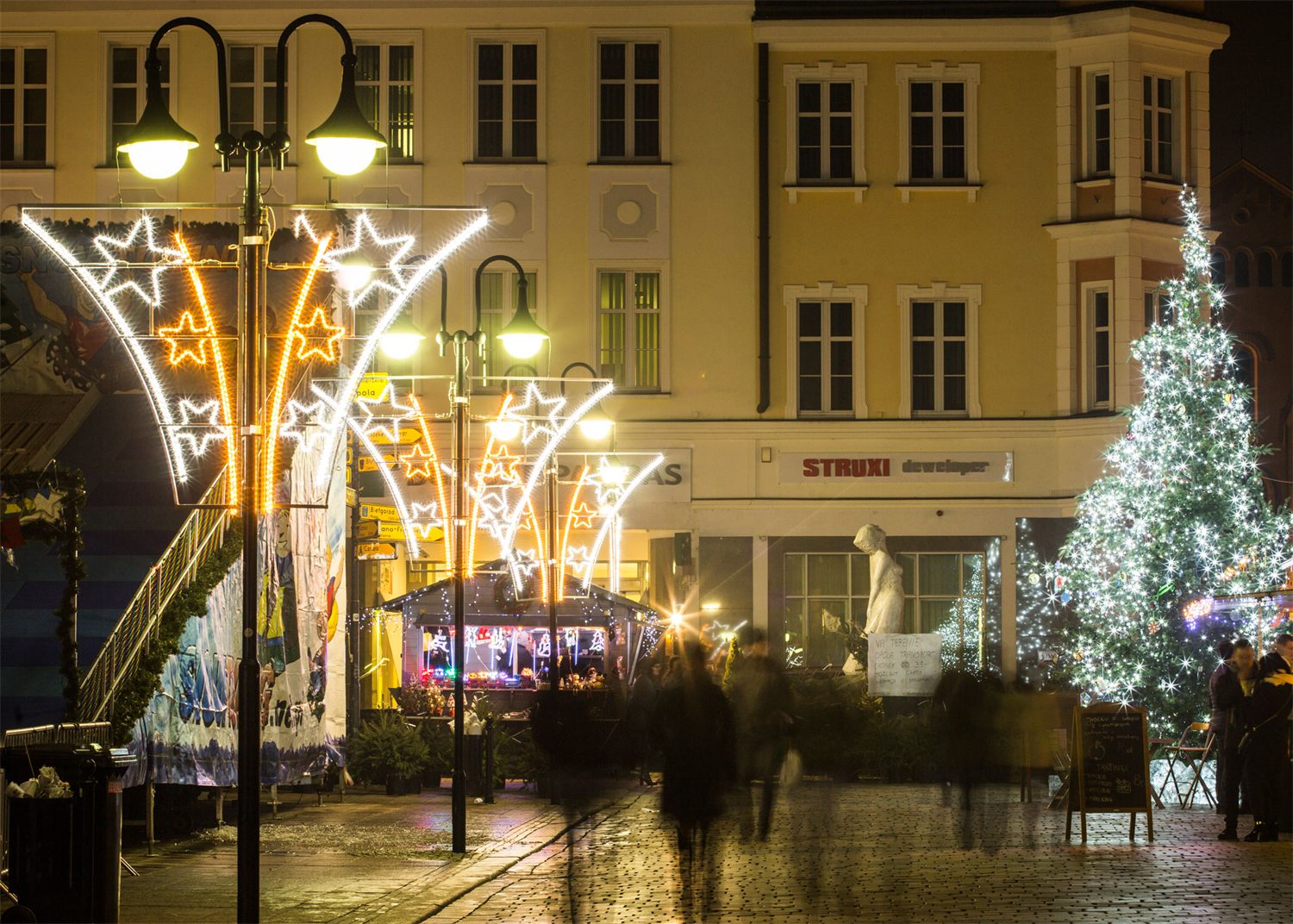 Gouden lichtornamenten aan de lantaarnpalen op een stadsplein met verlichte kerstboom